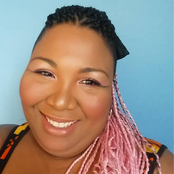 A Black woman with long braided ombre pink hair smiles at the camera.