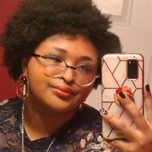 A Black and Indigenous person wearing glasses and red lipstick takes a selfie with his phone.