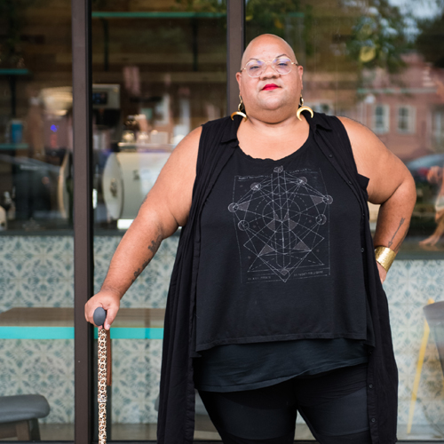 A Black non-binary person stands outside a cafe with their leopard print cane.