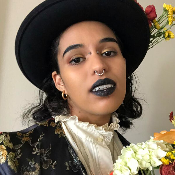 A Black person with black lipstick and pointed modified teeth looks at the camera while holding a bouquet.