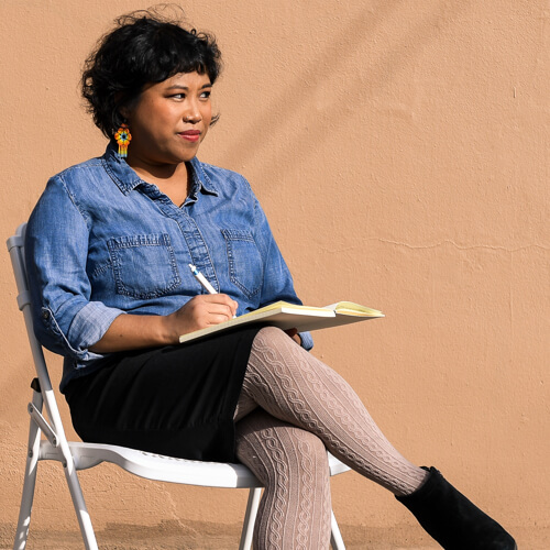 A Filipinx woman sits and pauses while writing in her notebook.