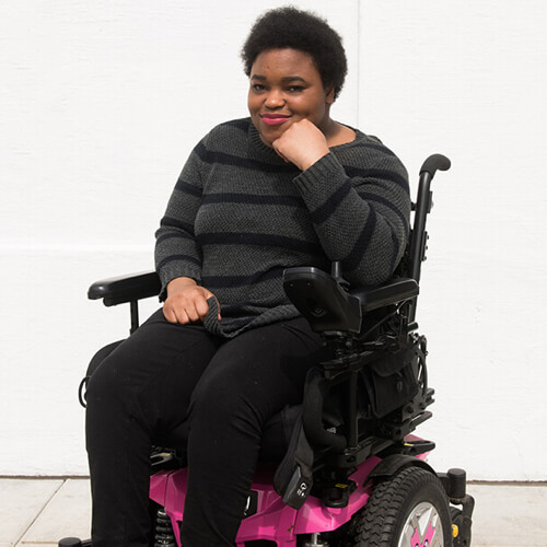 A Black non-binary person in a black and pink power wheelchair.