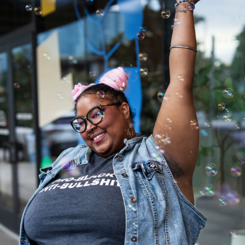 A Black person with glasses and pink buns smiles mid-twirl, surrounded by bubbles outdoors.