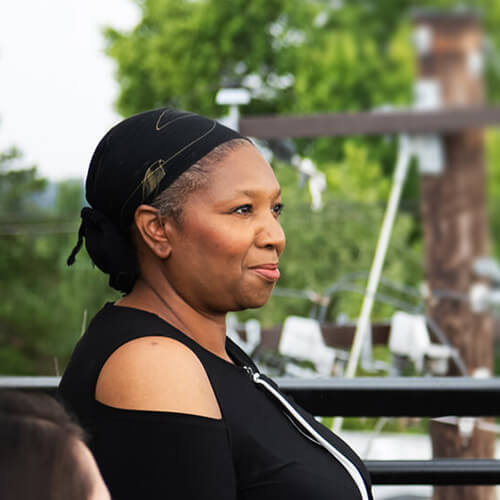 A Black woman wearing a head scarf sits on a rooftop deck, smiling and looking off to the side.
