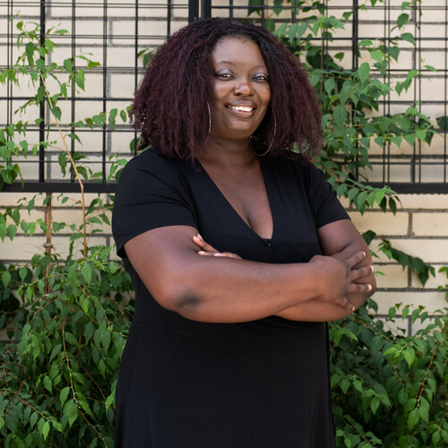 A Black woman smiles and has her arms crossed in front of a foliage wall.