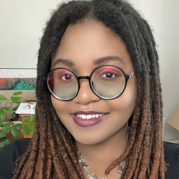 A Black person with long locs and round glasses smiling.